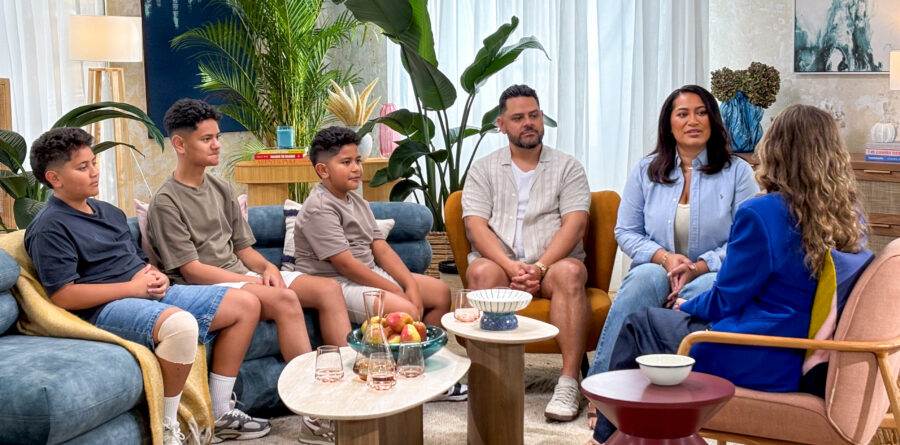 A family, comprised of two adults and three children sit on arm chairs with host, Ali Daddo